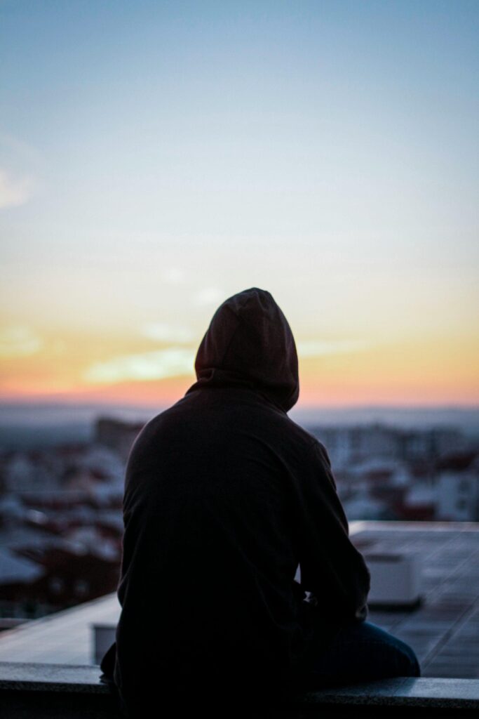 A solitary figure in a hoodie gazes at a vibrant sunset from a rooftop, creating a peaceful silhouette.